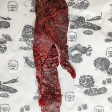 A long, thin piece of dark red cured meat lies on white paper printed with black illustrations of various meats, vegetables, and the label "Catoctin Lark Cured.