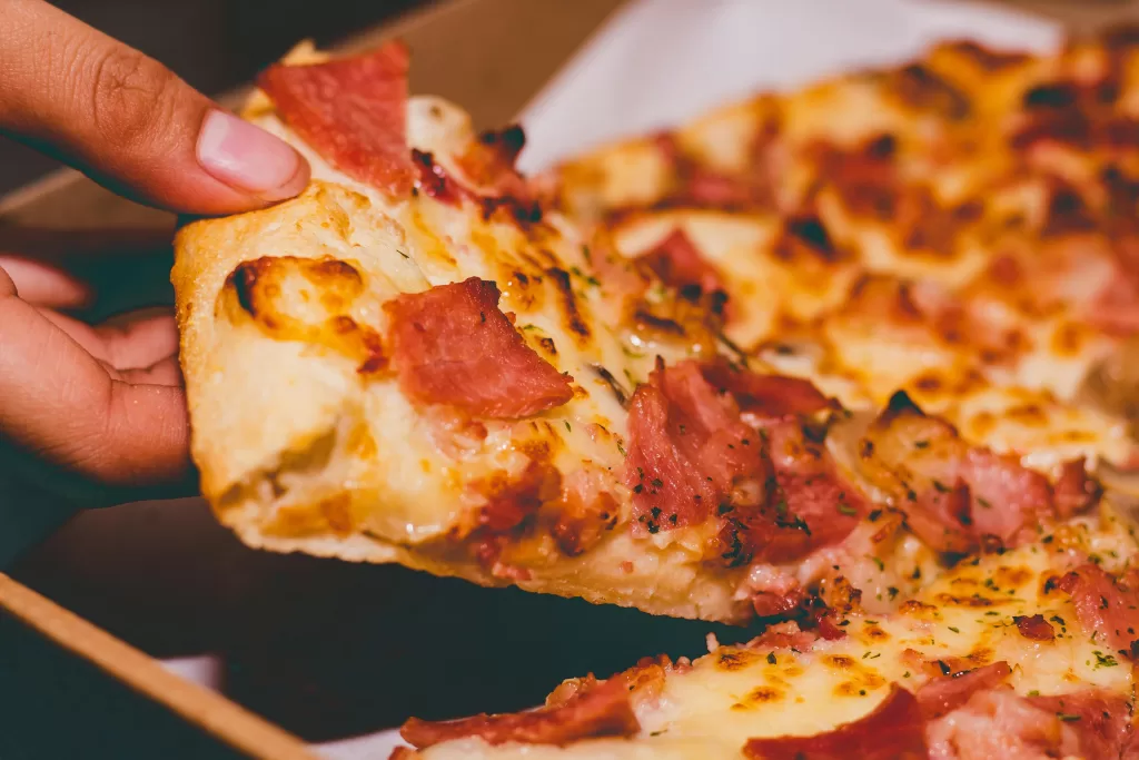 A close-up of a hand picking up a cheesy pizza slice topped with ham from a pizza box, perfect for celebrating national food holidays, with more slices visible in the background.