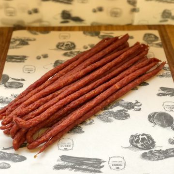 A bunch of thin, reddish-brown smoked meat sticks arranged in a neat pile on white deli paper with black illustrations of meats and the words "Cured".