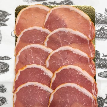 A package of thinly sliced cured meat, neatly arranged in overlapping rows on a gold tray, with a white paper featuring food illustrations and a label that reads “CURed.”.