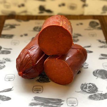 Three pieces of cured sausage, two stacked on the bottom and one balanced on top, displayed on illustrated butcher paper with meat-themed drawings and "Cured" labels in the background.