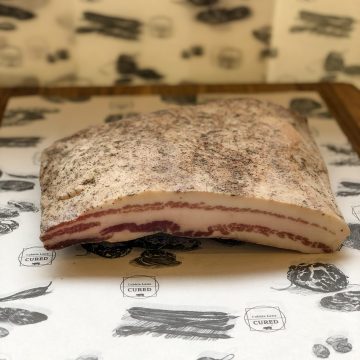 A slab of cured pork fat with a layer of meat, seasoned with spices, rests on white paper printed with "Cobb's Lane Cured" and illustrated meats. The background features similar printed designs.