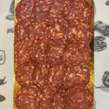 A golden tray holds neatly arranged slices of cured meat, likely salami, with visible bits of fat. The tray is placed on paper featuring black-and-white illustrations of meats and the words "Cobble Lane Cured.