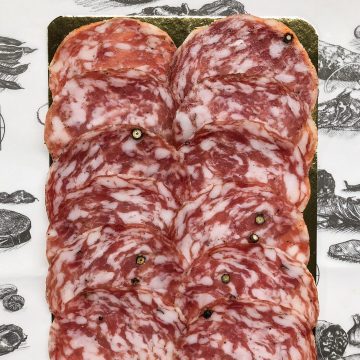 Thin slices of marbled salami are neatly arranged on a gold tray, with visible specks of black peppercorn. The tray is placed on paper featuring black and white illustrations of meats and a label that reads "Cobble Lane Cured.