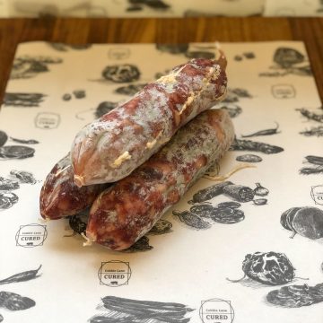 Two Islington Saucisson dry-cured sausages with a white, powdery coating are stacked on illustrated paper reading "Cobbles Lane Cured," displayed atop a wooden surface.