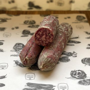 Three pieces of cured salami, one sliced to show the marbled interior, are arranged on butcher paper with food illustrations and "Cobble Lane Cured" logos in the background.