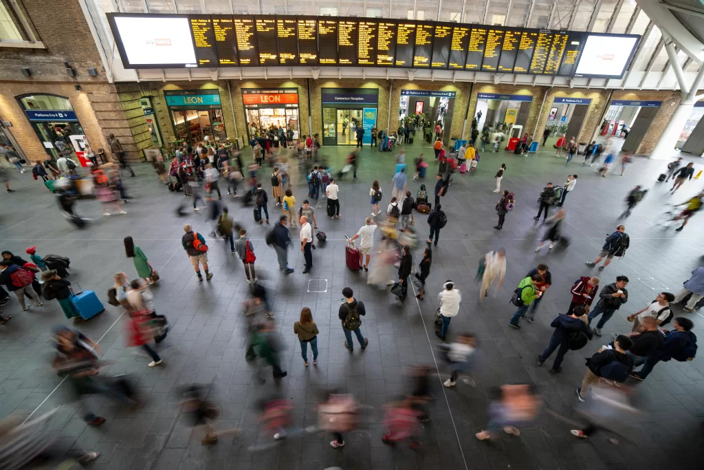 A busy train station with people walking and standing, some with luggage, beneath a large electronic departure board displaying train times and destinations. Among the lively crowd, an ad for London Restaurant Group catches travelers’ attention.