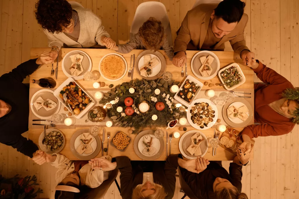 A group of people sit around a wooden table, holding hands in prayer before a festive meal. In this London Restaurant Group setting, candles, greenery, and various dishes create a warm and cozy atmosphere.
