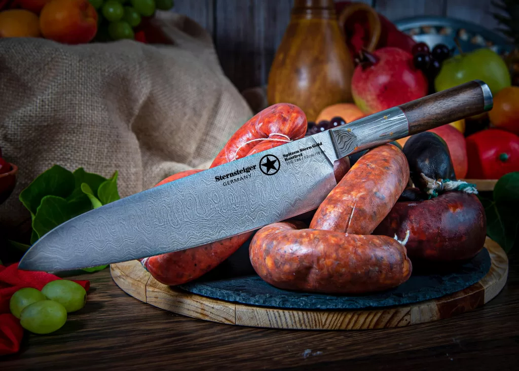 A chef’s knife with a patterned blade rests on several sausages on a round wooden board, surrounded by assorted fruits and vegetables—a scene inspired by the rustic charm of a London Restaurant Group kitchen.