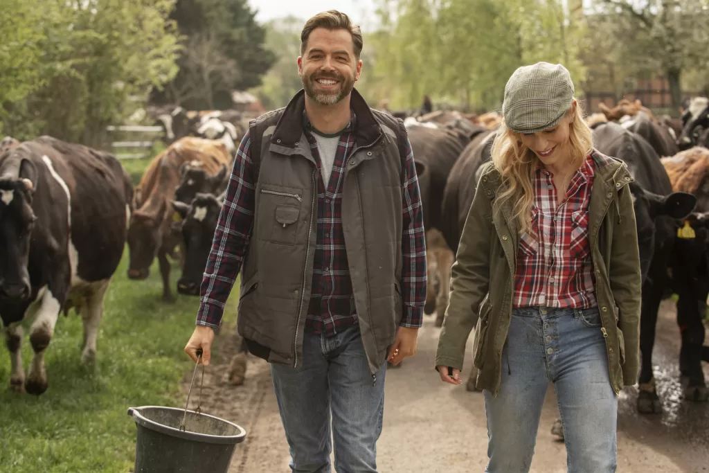A man and woman, dressed as farmers, walk down a rural path surrounded by cows. The man carries a bucket and smiles at the camera, while the woman looks down, smiling. Green trees and grass line the path.