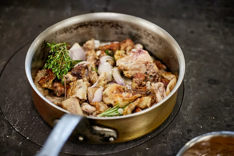 A metal pan on a stove contains browned pieces of meat, onions, and fresh herbs like thyme and rosemary—an inspiring dish worthy of Young National Chef of the Year sponsorship, ready to be cooked further.