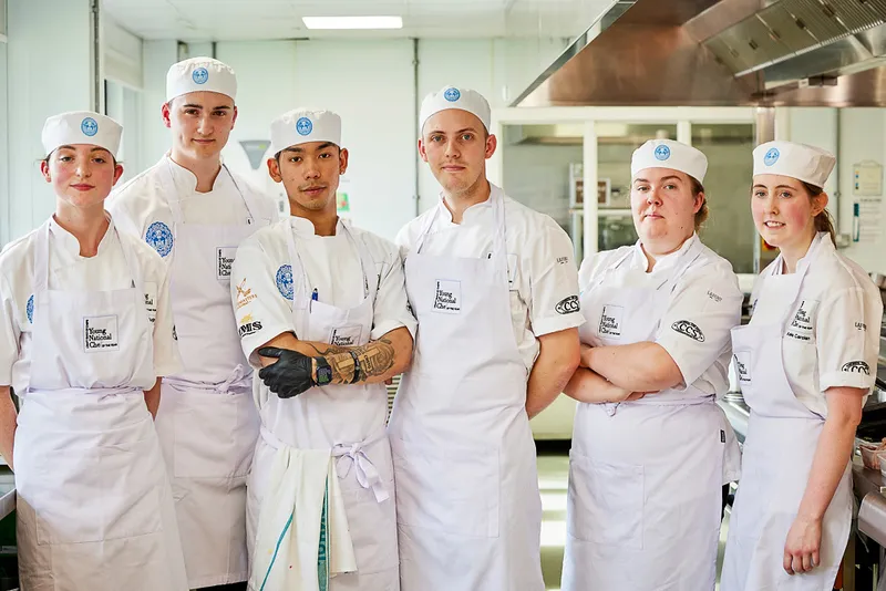 Six chefs in white uniforms and hats stand together in a professional kitchen, smiling proudly. Their camaraderie shines as they celebrate their Young National Chef of the Year sponsorship amidst gleaming stainless steel equipment.