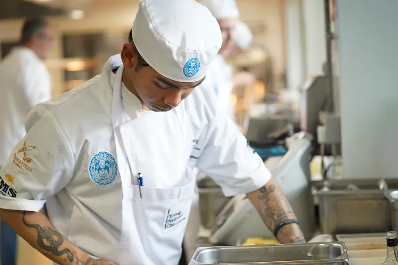 A chef in a white uniform and hat concentrates while preparing food in a professional kitchen, showcasing the skill and dedication inspired by Young National Chef of the Year sponsorship, with kitchen equipment and fellow chefs in the background.